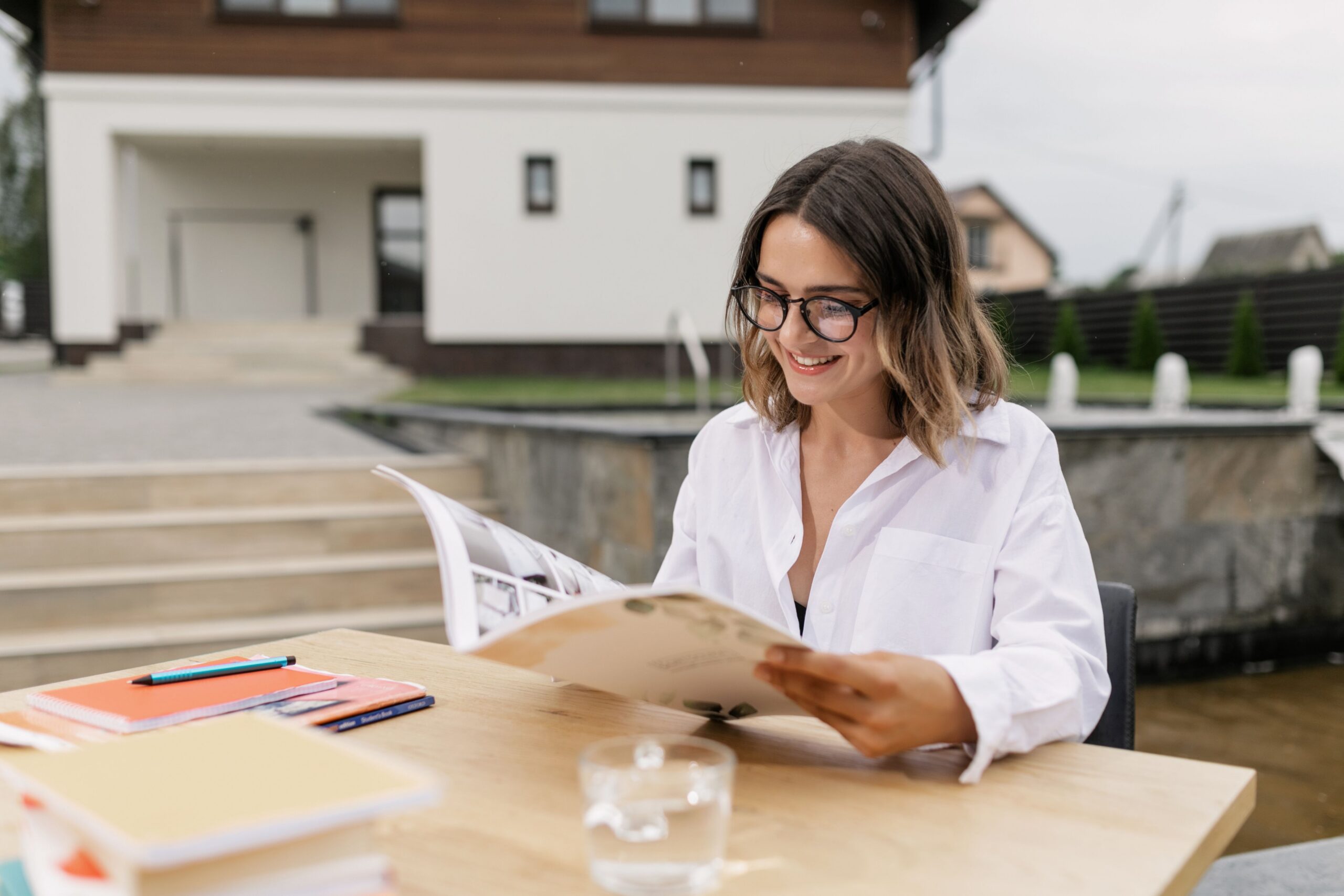 Woman in glasses reading a magazine outside at a table on a sunny day.